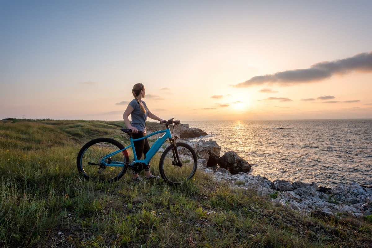 Woman with a bike in the nature / Morning view of a woman with an electric bike enjoys the view of sunrise at the rocky Black Sea coast